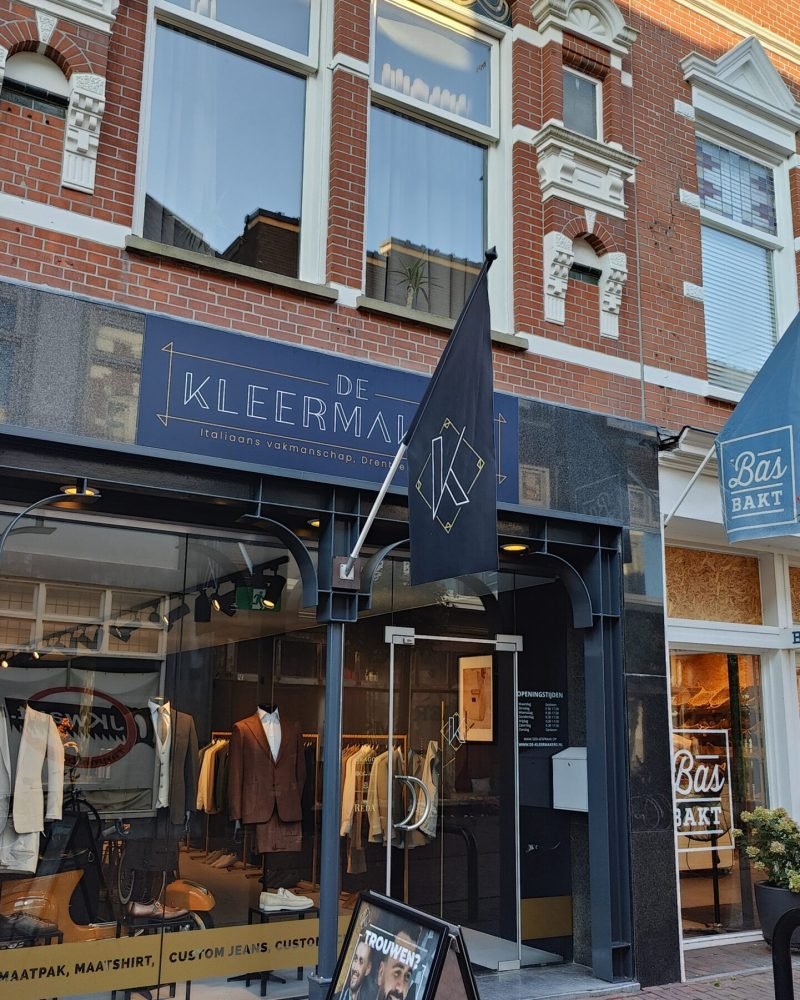 De gevel van de winkel De Kleermakers. Buiten staat een stoepbord en er hangt een vlag aan de gevel.