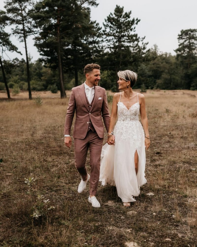 "Bruid en bruidegom wandelen hand in hand door een natuurgebied, gekleed in een roze trouwpak en een witte trouwjurk, terwijl ze elkaar liefdevol aankijken."